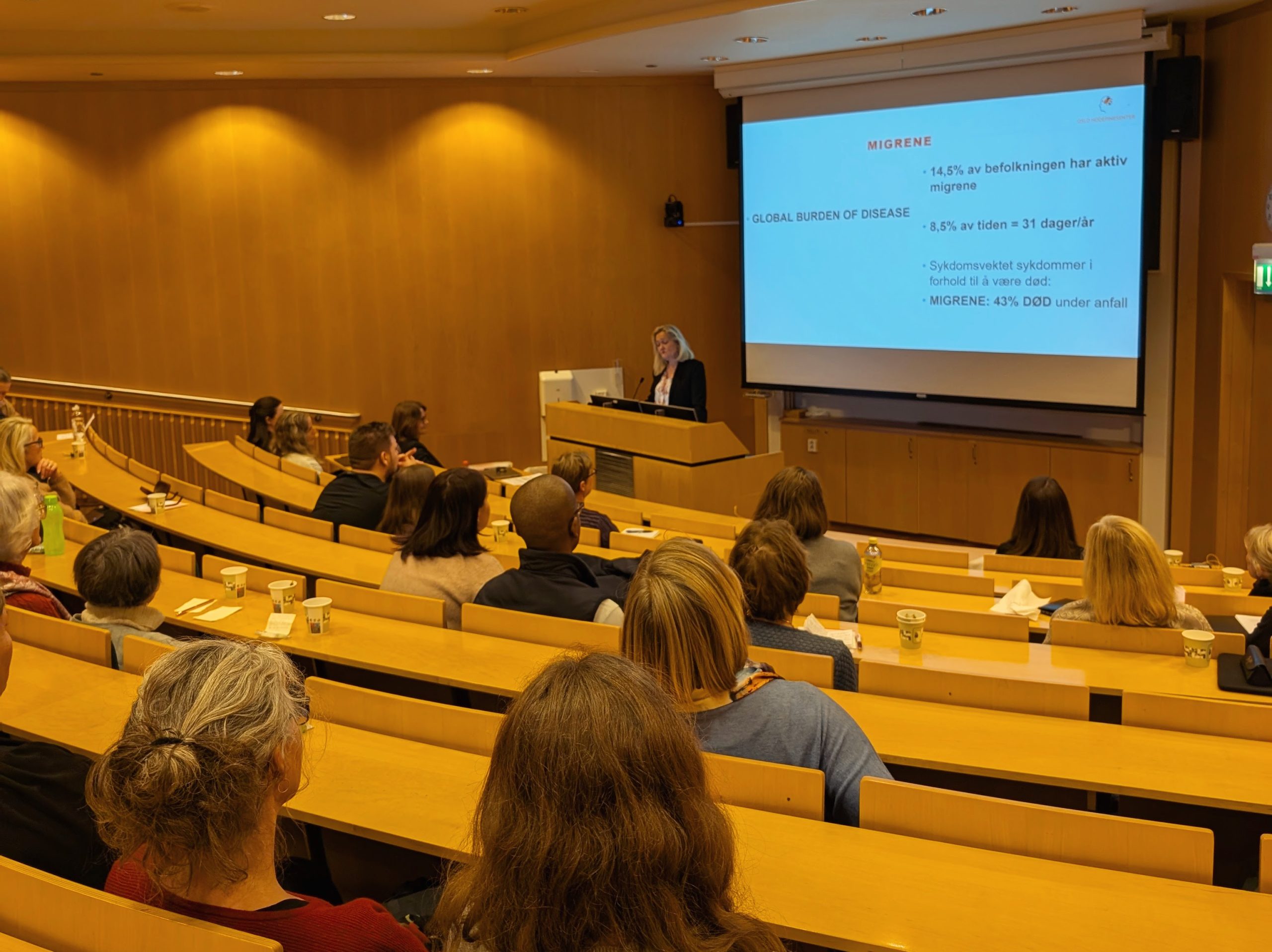 Bilde fra bakerst i grønt auditorie på Rikshospitalet. Vi ser bakhodene på publikum, og styreleder i Hodepine Norge som foreleser om migrene på scenen.