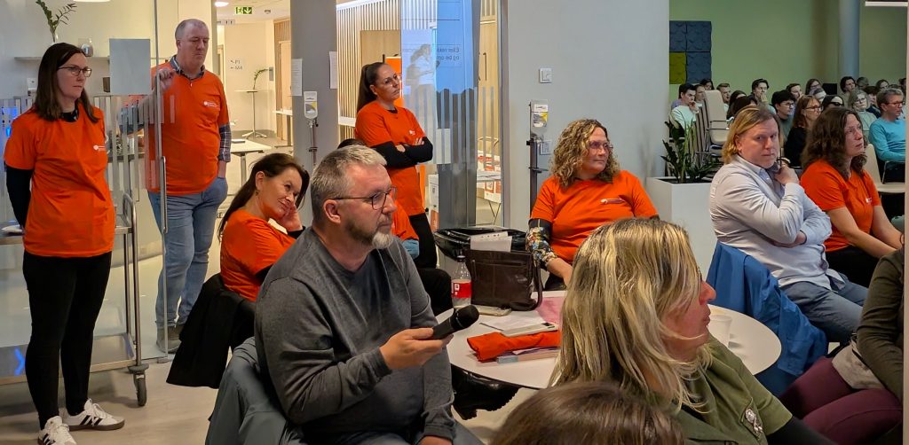 Flere representanter for Hodepine Norge Vestland i oransje T-skjorter, som står og sitter bakerst i lokalet og ser mot scenen. 