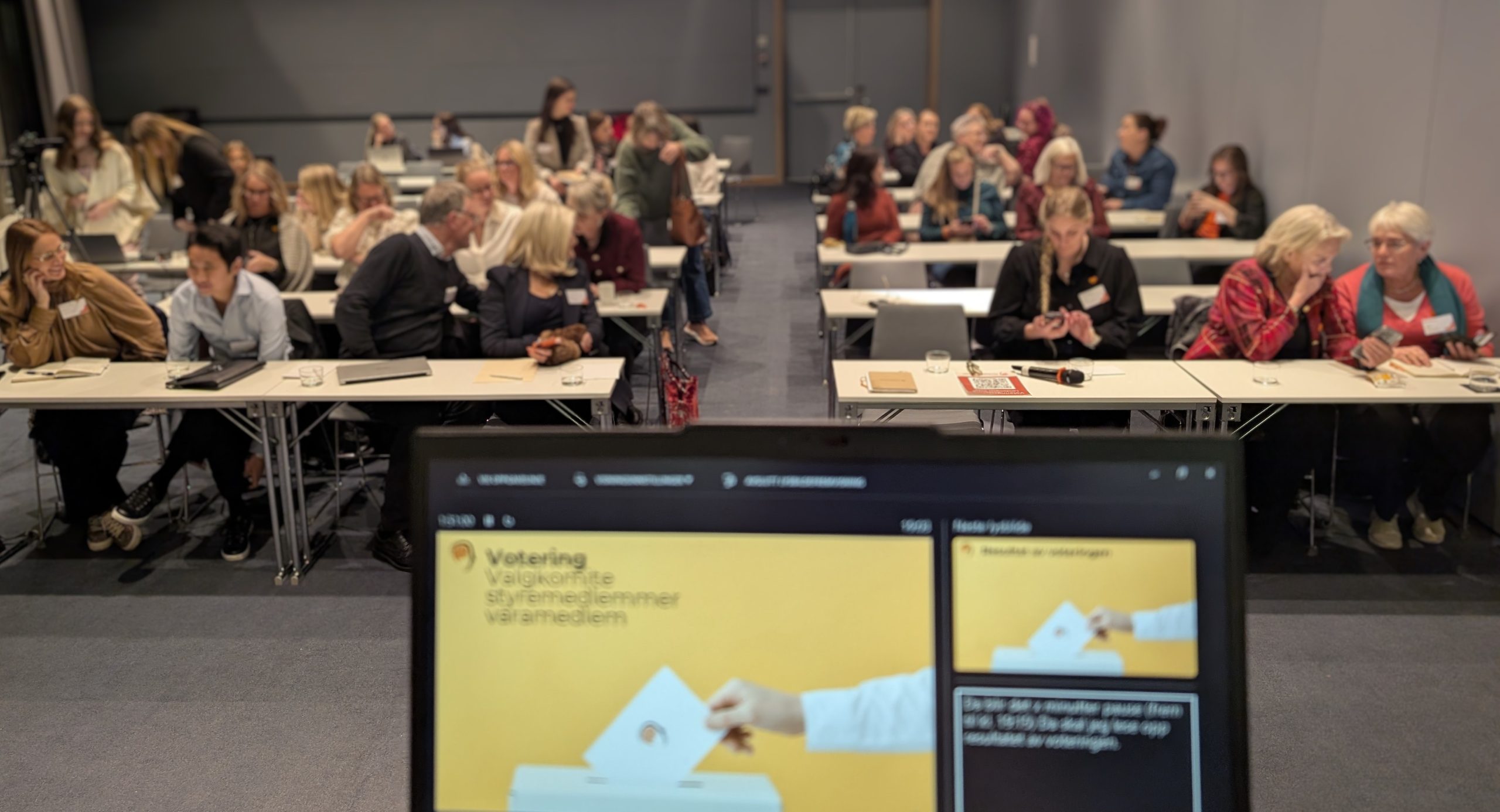 Hodepine Norges medlemmer stemmer under landsmøtet 2025. bildet er tatt fra scenen og viser en dataskjerm med teksten: Votering. I salen ser vi alle de fremmøtte. 